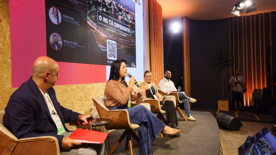 A imagem mostra um painel de discuss&atilde;o durante o evento South Summit Brazil. Quatro palestrantes est&atilde;o sentados em poltronas dispostas em semic&iacute;rculo sobre um palco elevado, de frente para uma plateia que n&atilde;o aparece na foto. Ao fundo, um grande tel&atilde;o exibe informa&ccedil;&otilde;es sobre a apresenta&ccedil;&atilde;o.

Detalhes do Palco e Palestrantes
Palestras: Tr&ecirc;s mulheres e um homem ocupam o palco. No centro da composi&ccedil;&atilde;o, uma mulher com cabelos longos e escuros, vestindo uma blusa rosa e cal&ccedil;a jeans pantalona, fala ao microfone. &Agrave; esquerda dela, um homem de terno azul segura uma pasta vermelha. &Agrave; direita, outra mulher e um homem observam a fala.

Mobili&aacute;rio: Os participantes est&atilde;o sentados em poltronas confort&aacute;veis de cor bege com p&eacute;s de madeira. Pequenas mesas de apoio redondas est&atilde;o posicionadas entre as poltronas.

O Tel&atilde;o: Exibe a frase em destaque "O RS T&Aacute; DIFERENTE" sobre uma foto a&eacute;rea noturna de uma cidade. No lado esquerdo do tel&atilde;o, aparecem fotos e nomes dos painelistas: Danielle Calazans, Adriane Perin e Nielson Carrancilo. H&aacute; tamb&eacute;m um QR Code grande na parte inferior central.

Cen&aacute;rio e Ilumina&ccedil;&atilde;o
Ambiente: O palco tem um acabamento r&uacute;stico e moderno, com paredes de placas de madeira (OSB) e detalhes em ripas de madeira verticais ao fundo. O ch&atilde;o do palco &eacute; coberto por um tapete cinza escuro.

Ilumina&ccedil;&atilde;o: Refletores de palco est&atilde;o vis&iacute;veis no teto &agrave; direita, lan&ccedil;ando uma luz quente sobre os palestrantes. No canto inferior direito, um monitor de retorno no ch&atilde;o exibe a mesma imagem do tel&atilde;o principal.