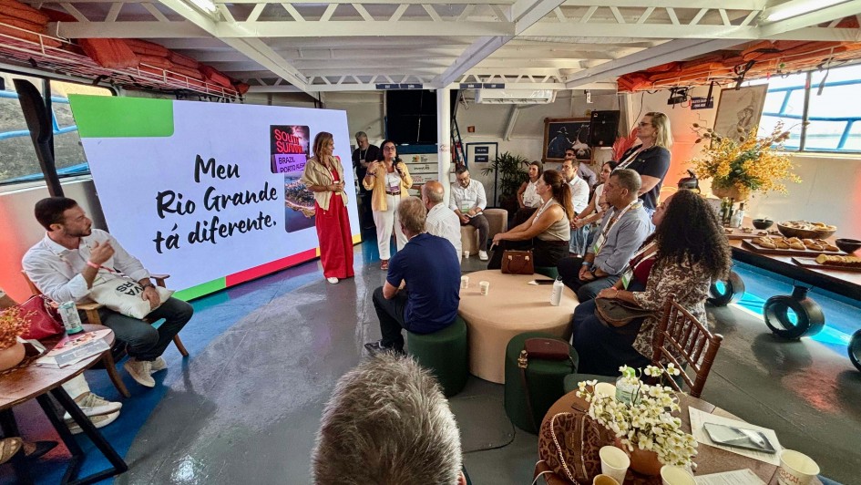 A imagem mostra um encontro realizado em um espa&ccedil;o interno iluminado por luz natural lateral. Um tel&atilde;o grande exibe a frase &ldquo;Meu Rio Grande t&aacute; diferente.&rdquo;, acompanhada da identidade visual do South Summit Brazil. Pessoas est&atilde;o distribu&iacute;das em assentos baixos e mesas de apoio, enquanto duas pessoas ficam &agrave; frente do tel&atilde;o conduzindo a apresenta&ccedil;&atilde;o.