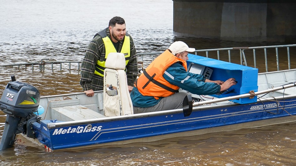 Imagem mostra dois homens num pequeno barco, no meio de um rio grande, pr&oacute;ximo a um pilar de uma ponte. Eles est&atilde;o com equipamentos de batimetria para medi&ccedil;&atilde;o do leito do rio.