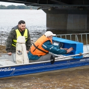Imagem mostra dois homens num pequeno barco, no meio de um rio grande, pr&oacute;ximo a um pilar de uma ponte. Eles est&atilde;o com equipamentos de batimetria para medi&ccedil;&atilde;o do leito do rio.