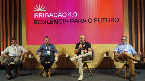 Painel realizado em palco de evento, com quatro participantes sentados em poltronas individuais, cada um com mesa lateral e copo de apoio. Ao fundo, um grande tel&atilde;o exibe o t&iacute;tulo &ldquo;IRRIGA&Ccedil;&Atilde;O 4.0: RESILI&Ecirc;NCIA PARA O FUTURO&rdquo;, com identidade visual em tons de vermelho e laranja.
