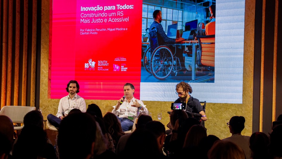 Foto mostra tr&ecirc;s palestrantes no palco, sentado. Um deles &eacute; cadeirante. Eles falam diante da plateia. Atr&aacute;s, h&aacute; uma tela com o nome da palestra e a foto de um cadeirante, com terno, diante de uma mesa de computador em um ambiente de escrit&oacute;rio. 