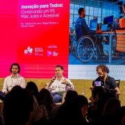 Foto mostra tr&ecirc;s palestrantes no palco, sentado. Um deles &eacute; cadeirante. Eles falam diante da plateia. Atr&aacute;s, h&aacute; uma tela com o nome da palestra e a foto de um cadeirante, com terno, diante de uma mesa de computador em um ambiente de escrit&oacute;rio. 
