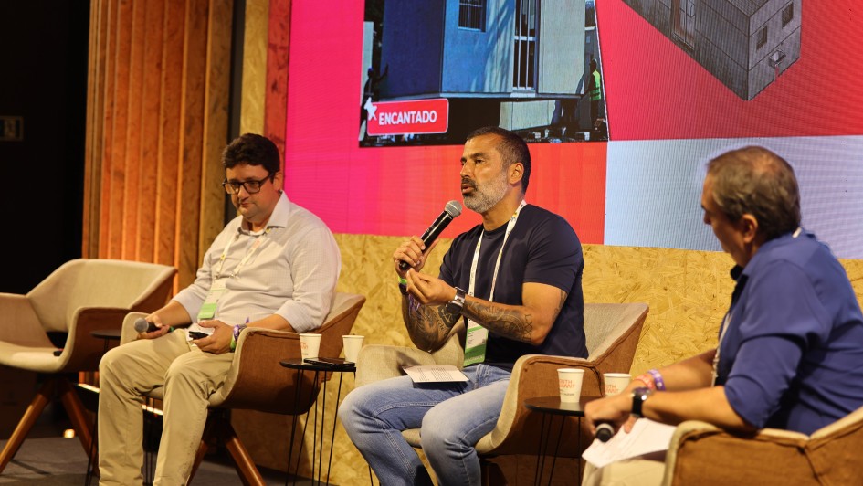 A imagem mostra um painel em palco de evento, com tr&ecirc;s participantes sentados em poltronas individuais, cada um com mesa lateral. Uma das pessoas utiliza um microfone durante a fala.
