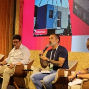 A imagem mostra um painel em palco de evento, com tr&ecirc;s participantes sentados em poltronas individuais, cada um com mesa lateral. Uma das pessoas utiliza um microfone durante a fala.