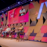 Esta &eacute; uma descri&ccedil;&atilde;o detalhada da imagem para fins de acessibilidade:

Vis&atilde;o Geral
A imagem mostra um painel de debate no evento South Summit Brazil. Quatro homens est&atilde;o sentados em banquetas altas de madeira sobre um palco, participando de uma discuss&atilde;o. Ao fundo, um cen&aacute;rio din&acirc;mico e um grande tel&atilde;o comp&otilde;em o ambiente.

Detalhes do Palco e Palestrantes
Os Participantes: Da esquerda para a direita, o primeiro homem veste uma camisa polo clara e cal&ccedil;a bege; o segundo, uma camisa social azul clara e jeans; o terceiro, uma camiseta preta; e o quarto, que est&aacute; falando e gesticulando no momento, veste uma camisa polo bege e cal&ccedil;a escura. Todos utilizam crach&aacute;s do evento.

O Tel&atilde;o: Exibe uma transmiss&atilde;o ao vivo em close-up do palestrante que est&aacute; falando. No lado esquerdo do tel&atilde;o, h&aacute; um texto em ingl&ecirc;s sobre fundo rosa: "BLENDED FINANCE FOR RESILIENT DEVELOPMENT: THE ROLE OF DEVELOPMENT BANKS AND CATALYTIC PHILANTHROPY". Abaixo, os nomes dos participantes e suas institui&ccedil;&otilde;es s&atilde;o listados em letras menores.

Cen&aacute;rio de Fundo: A parede atr&aacute;s dos palestrantes &eacute; decorada com pain&eacute;is geom&eacute;tricos sobrepostos em tons de madeira natural, rosa, azul escuro e cinza. Algumas pe&ccedil;as possuem o logotipo do evento e do Governo do Estado do Rio Grande do Sul. Uma grande estrutura luminosa em formato de "V" invertido ou parte de um "A" se destaca no lado direito.

Composi&ccedil;&atilde;o e Ilumina&ccedil;&atilde;o
Ambiente: O palco tem um acabamento em carpete bege claro. Uma fileira de luzes LED no ch&atilde;o, na borda frontal do palco, emite uma luz azulada. No topo da estrutura, refletores de palco iluminam a cena.

Perspectiva: A foto &eacute; tirada de um &acirc;ngulo lateral e ligeiramente de baixo, capturando a extens&atilde;o do palco e a modernidade da cenografia.
