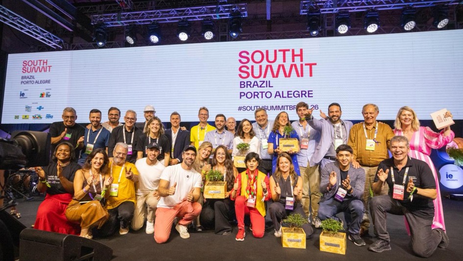 Registro coletivo no palco do South Summit Brazil &ndash; Porto Alegre, com um grupo numeroso reunido para premia&ccedil;&atilde;o. As pessoas est&atilde;o posicionadas em p&eacute; e agachadas, algumas segurando caixas de madeira com plantas. Ao fundo, um grande tel&atilde;o exibe a identidade visual do evento, com ilumina&ccedil;&atilde;o c&ecirc;nica e estrutura profissional de palco.