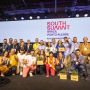 Registro coletivo no palco do South Summit Brazil &ndash; Porto Alegre, com um grupo numeroso reunido para premia&ccedil;&atilde;o. As pessoas est&atilde;o posicionadas em p&eacute; e agachadas, algumas segurando caixas de madeira com plantas. Ao fundo, um grande tel&atilde;o exibe a identidade visual do evento, com ilumina&ccedil;&atilde;o c&ecirc;nica e estrutura profissional de palco.
