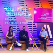 Esta &eacute; uma descri&ccedil;&atilde;o detalhada da imagem para fins de acessibilidade:

Vis&atilde;o Geral
A imagem apresenta um painel de debate no evento South Summit Brazil 2026. Tr&ecirc;s palestrantes (uma mulher e dois homens) est&atilde;o sentados em poltronas modernas sobre um palco, engajados em uma conversa. O cen&aacute;rio ao fundo &eacute; vibrante e utiliza elementos de madeira e ilumina&ccedil;&atilde;o colorida.

Detalhes do Palco e Palestrantes
Os Participantes: &Agrave; esquerda, uma mulher de cabelos escuros e &oacute;culos, vestindo um vestido marrom e t&ecirc;nis brancos, observa o colega. No centro, um homem de barba, usando um blazer escuro, camiseta preta e t&ecirc;nis brancos, segura um microfone. &Agrave; direita, um homem de camisa social azul clara e cal&ccedil;a escura fala ao microfone, gesticulando com a m&atilde;o esquerda. Todos portam crach&aacute;s oficiais do evento.

Mobili&aacute;rio: Os palestrantes ocupam poltronas girat&oacute;rias de cor ferrugem/terrosa com base cil&iacute;ndrica. Entre eles, h&aacute; pequenas mesas de apoio circulares de madeira clara.

Cen&aacute;rio de Fundo: O fundo &eacute; composto por pain&eacute;is de madeira ripada (estilo pallet) sobrepostos por grandes faixas diagonais transl&uacute;cidas em tons de rosa e roxo. No centro, destaca-se a inscri&ccedil;&atilde;o em relevo prateado: "SOUTH SUMMIT BRAZIL PORTO ALEGRE".

Elementos Digitais e Ilumina&ccedil;&atilde;o
Monitores: Dois monitores laterais exibem informa&ccedil;&otilde;es do painel. O da direita mostra o t&iacute;tulo em ingl&ecirc;s sobre um fundo magenta: "HEALTH IN TRANSITION: TRANSFORMING SYSTEM PRESSURES INTO INNOVATION AND NEW MODELS OF VALUE", al&eacute;m dos nomes dos moderadores e participantes.

Composi&ccedil;&atilde;o: A ilumina&ccedil;&atilde;o &eacute; difusa e colorida, com tons de azul e rosa predominando no ambiente. O palco tem um revestimento claro e neutro. &Agrave; extrema direita, &eacute; vis&iacute;vel parte de uma estrutura de treli&ccedil;a met&aacute;lica (box truss) comum em grandes eventos.