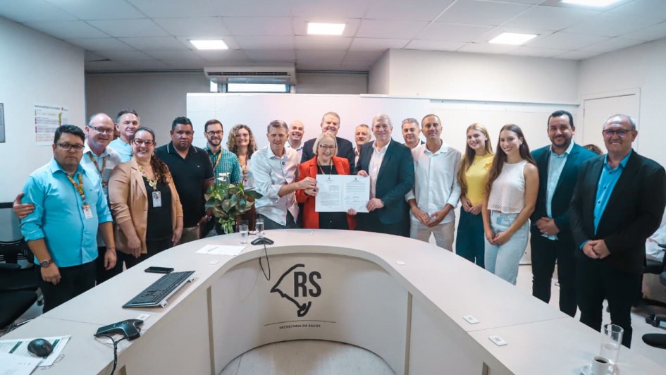 A imagem mostra uma reuni&atilde;o institucional em uma sala de reuni&otilde;es ampla, com mesa em formato semicircular. Um grupo numeroso est&aacute; reunido em p&eacute; atr&aacute;s da mesa, enquanto, ao centro, duas pessoas seguram um documento aberto.