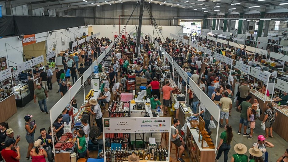 A imagem mostra uma feira agroindustrial realizada em um grande pavilh&atilde;o coberto, com corredores longos e estandes organizados em fileiras paralelas. Os estandes exibem produtos aliment&iacute;cios, bebidas, utens&iacute;lios e itens artesanais, com bancadas de madeira e sinaliza&ccedil;&atilde;o suspensa indicando marcas e expositores. O espa&ccedil;o est&aacute; intensamente ocupado por visitantes circulando pelos corredores. 