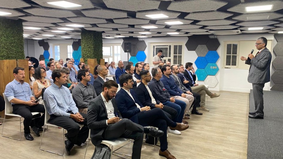 Imagem mostra uma sala com v&aacute;rias pessoas sentadas e uma delas &agrave; frente deles de p&eacute; e com microfone