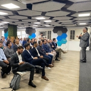 Imagem mostra uma sala com v&aacute;rias pessoas sentadas e uma delas &agrave; frente deles de p&eacute; e com microfone