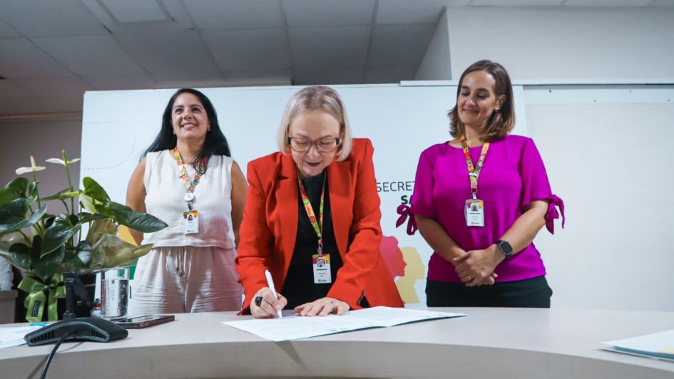 A imagem mostra a secret&aacute;ria da Sa&uacute;de assinando um documento sobre uma mesa clara em ambiente institucional, enquanto duas outras pessoas permanecem ao fundo observando.