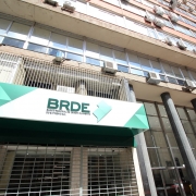 Vis&atilde;o Geral
A imagem mostra a fachada de um edif&iacute;cio comercial de m&uacute;ltiplos andares, com foco em uma placa de identifica&ccedil;&atilde;o do BRDE (Banco Regional de Desenvolvimento do Extremo Sul). A foto &eacute; tirada de um &acirc;ngulo de baixo para cima (contra-plong&eacute;e), enfatizando a altura da estrutura.

Detalhes da Fachada e Identifica&ccedil;&atilde;o
Placa do Banco: Em primeiro plano, no lado esquerdo, h&aacute; um painel horizontal moderno com fundo branco e detalhes geom&eacute;tricos em tons de verde. Nele, l&ecirc;-se em letras escuras: "BRDE - BANCO REGIONAL DE DESENVOLVIMENTO DO EXTREMO SUL", acompanhado por um logotipo verde que remete ao mapa da regi&atilde;o Sul do Brasil.

O Edif&iacute;cio: A estrutura possui uma arquitetura funcional, com janelas de vidro que refletem a luz do dia e diversas unidades de ar-condicionado fixadas na parede externa de tijolos aparentes. Colunas cil&iacute;ndricas escuras sustentam a parte frontal do pr&eacute;dio no n&iacute;vel da cal&ccedil;ada.

Entrada: Abaixo da placa do BRDE, v&ecirc;-se uma grade met&aacute;lica de prote&ccedil;&atilde;o e uma porta de vidro que d&aacute; acesso ao interior do banco.

Composi&ccedil;&atilde;o e Ilumina&ccedil;&atilde;o
Perspectiva: O &acirc;ngulo acentuado cria uma linha diagonal que guia o olhar da placa no canto inferior esquerdo at&eacute; o topo do edif&iacute;cio no canto superior direito.

Ilumina&ccedil;&atilde;o: A luz solar &eacute; direta e clara, indicando um dia ensolarado. O c&eacute;u azul &eacute; vis&iacute;vel no canto superior direito, contrastando com os tons de cinza, bege e verde da edifica&ccedil;&atilde;o.