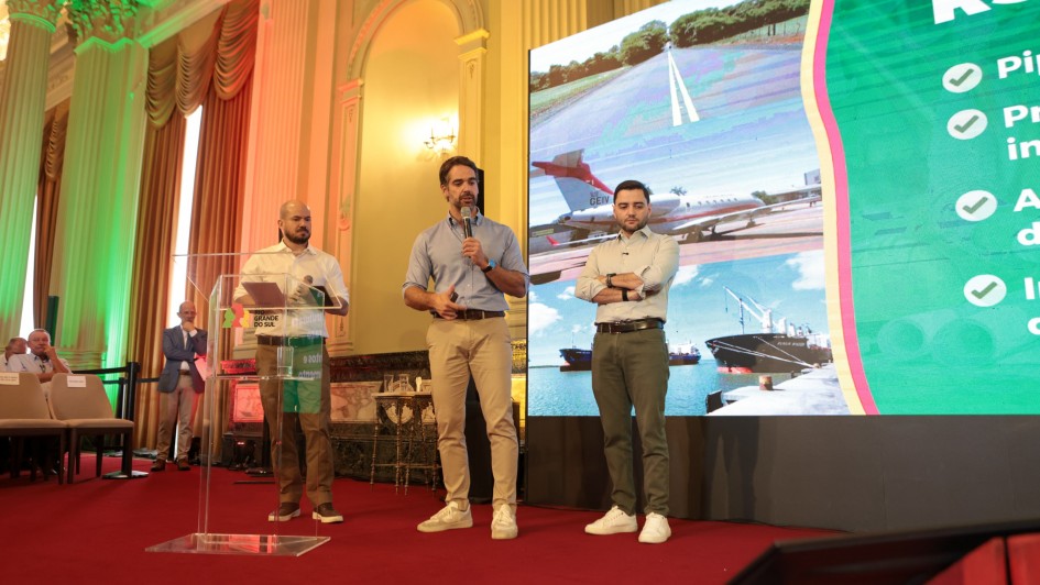Tr&ecirc;s homens est&atilde;o em um palco com tapete vermelho, um deles fala ao microfone, enquanto outro observa e o terceiro tamb&eacute;m segura um microfone. Ao fundo, uma grande tela mostrando estrada, avi&atilde;o e navios.