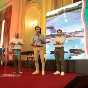 Tr&ecirc;s homens est&atilde;o em um palco com tapete vermelho, um deles fala ao microfone, enquanto outro observa e o terceiro tamb&eacute;m segura um microfone. Ao fundo, uma grande tela mostrando estrada, avi&atilde;o e navios.