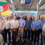 Grupo de cerca de 15 homens posando para foto dentro da Esta&ccedil;&atilde;o Rodovi&aacute;ria de Porto Alegre. Ao fundo, a bandeira do Rio Grande do Sul e um painel com logotipos institucionais do governo estadual. O ambiente &eacute; coberto, com estrutura de concreto aparente. As placas indicam &aacute;reas como "Embarque interestadual e internacional" e "Sala de Embarque".