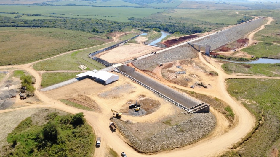 Foto a&eacute;rea mostra extens&atilde;o da obra da barragem, ainda sem &aacute;gua. H&aacute; trechos j&aacute; constru&iacute;dos, outros que est&atilde;o aterrados e h&aacute;, ao fundo, &aacute;rea verde de campo e um rio que corta a regi&atilde;o.