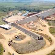 Foto a&eacute;rea mostra extens&atilde;o da obra da barragem, ainda sem &aacute;gua. H&aacute; trechos j&aacute; constru&iacute;dos, outros que est&atilde;o aterrados e h&aacute;, ao fundo, &aacute;rea verde de campo e um rio que corta a regi&atilde;o.