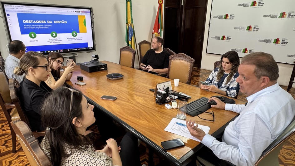 A imagem mostra uma reuni&atilde;o de trabalho em uma sala de reuni&otilde;es de estilo cl&aacute;ssico. Sete pessoas est&atilde;o sentadas ao redor de uma grande mesa de madeira retangular, participando de uma apresenta&ccedil;&atilde;o institucional. Um homem em destaque &agrave; direita fala enquanto opera um computador.

Detalhes dos Participantes
O Palestrante: No canto inferior direito, um homem de pele clara e cabelos curtos grisalhos, vestindo uma camisa social azul clara, gesticula com a m&atilde;o esquerda enquanto a direita opera um mouse sobre a mesa.

O Grupo: Outras seis pessoas (quatro mulheres e dois homens) est&atilde;o distribu&iacute;das ao redor da mesa. Algumas observam o palestrante, enquanto outras olham para uma tela ou para seus pr&oacute;prios celulares. Eles vestem roupas casuais e profissionais.

Mobili&aacute;rio: A mesa &eacute; de madeira clara com bordas escuras. As cadeiras s&atilde;o de madeira trabalhada com estofado em tom de couro marrom.

Elementos de Apoio e Ambiente
Monitor Digital: No fundo, &agrave; esquerda, h&aacute; um grande monitor de tela plana montado em um suporte m&oacute;vel. A tela exibe um slide com o t&iacute;tulo "DESTAQUES DA GEST&Atilde;O" e tr&ecirc;s pontos numerados sobre um fundo azul e branco.

Decora&ccedil;&atilde;o e S&iacute;mbolos: Ao fundo, no centro, est&atilde;o posicionadas duas bandeiras em mastros: a bandeira do Brasil e a bandeira do Rio Grande do Sul. &Agrave; direita, h&aacute; um grande painel branco (backdrop) com logotipos repetidos da Secretaria da Agricultura, Pecu&aacute;ria, Produ&ccedil;&atilde;o Sustent&aacute;vel e Irriga&ccedil;&atilde;o do Governo do Estado do Rio Grande do Sul.

Arquitetura: O piso &eacute; de madeira com um padr&atilde;o geom&eacute;trico de marchetaria (parquet). As paredes s&atilde;o brancas e h&aacute; uma porta de madeira escura ao fundo. Sobre a mesa, veem-se celulares, um telefone de confer&ecirc;ncia, um teclado preto, pap&eacute;is e copos de &aacute;gua.

Composi&ccedil;&atilde;o
A foto &eacute; tirada de um &acirc;ngulo superior lateral, capturando quase toda a extens&atilde;o da mesa e criando uma perspectiva que integra todos os participantes e os elementos institucionais da sala.