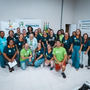 Esta &eacute; uma descri&ccedil;&atilde;o detalhada da imagem para fins de acessibilidade:

Vis&atilde;o Geral
A imagem mostra um grupo de aproximadamente 25 pessoas reunidas em um ambiente interno para uma fotografia coletiva. O grupo &eacute; composto majoritariamente por mulheres, muitas vestindo uniformes de uma institui&ccedil;&atilde;o. No centro, destaca-se uma autoridade governamental.

Detalhes do Grupo
Vestimentas: A maioria dos presentes usa camisetas de cor azul-petr&oacute;leo com golas laranjas ou camisetas verdes. Muitas dessas pe&ccedil;as exibem o logotipo da APAE Tapes.

Figura Central: No centro do grupo, est&aacute; uma mulher de pele clara, cabelos loiros curtos e &oacute;culos, vestindo um conjunto de blazer e cal&ccedil;a em tom azul-celeste claro. Ela sorri para a c&acirc;mera.

Disposi&ccedil;&atilde;o: As pessoas est&atilde;o organizadas em tr&ecirc;s n&iacute;veis: algumas ajoelhadas &agrave; frente, outras em p&eacute; no meio e uma fileira ao fundo. O clima &eacute; de celebra&ccedil;&atilde;o e uni&atilde;o.

Banners e Identifica&ccedil;&atilde;o
Ao fundo, tr&ecirc;s banners brancos fornecem o contexto do evento:

Banner &agrave; esquerda: Cont&eacute;m o logotipo colorido do programa TEAcolhe RS (Programa de Atendimento a Pessoas com Autismo).

Banner central: Exibe o logotipo da Secretaria de Educa&ccedil;&atilde;o do Governo do Estado do Rio Grande do Sul.

Banner &agrave; direita: Lista "Programas com foco na alfabetiza&ccedil;&atilde;o", mencionando iniciativas como o Compromisso Nacional Crian&ccedil;a Alfabetizada e o Programa Alfabetiza Tch&ecirc;.

Ambiente
Cen&aacute;rio: O local tem paredes brancas e ch&atilde;o de cer&acirc;mica clara. Ao fundo, entre os banners, s&atilde;o vis&iacute;veis as bandeiras do Brasil e do Rio Grande do Sul em mastros.

Composi&ccedil;&atilde;o: A ilumina&ccedil;&atilde;o &eacute; uniforme e direta. No canto inferior direito, &eacute; poss&iacute;vel ver parte de algumas bolsas e cadeiras, sugerindo que o grupo est&aacute; em um audit&oacute;rio ou sala de eventos multifuncional.