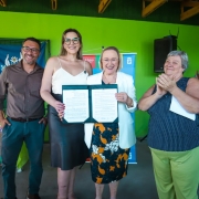 A imagem mostra um grupo reunido em frente a uma parede verde. No centro, duas pessoas seguram um documento aberto voltado para a c&acirc;mera. Ao fundo, h&aacute; banners institucionais e elementos de decora&ccedil;&atilde;o simples.