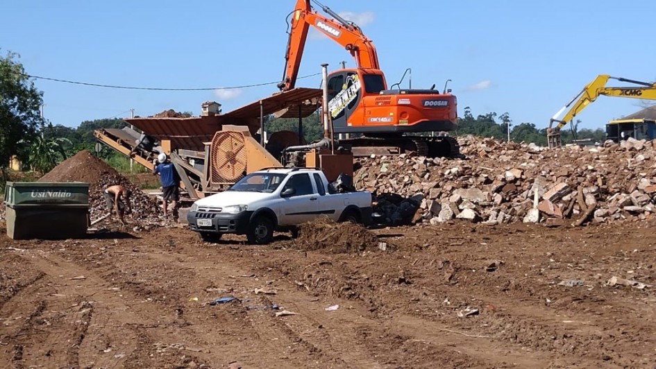 Processo de britagem de entulho, com escavadeira e m&aacute;quina de peneiramento operando sobre um grande volume de material demolido. Um ve&iacute;culo utilit&aacute;rio est&aacute; parado pr&oacute;ximo ao equipamento, e trabalhadores movimentam entulhos em meio ao solo revolvido.