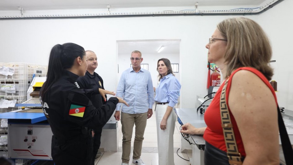 Visita em &aacute;rea de trabalho, com pessoas reunidas ao redor de m&aacute;quinas e bancadas. O espa&ccedil;o possui equipamentos de produ&ccedil;&atilde;o, caixas organizadoras, extintor de inc&ecirc;ndio fixado na parede e ilumina&ccedil;&atilde;o branca uniforme.