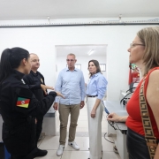 Visita em &aacute;rea de trabalho, com pessoas reunidas ao redor de m&aacute;quinas e bancadas. O espa&ccedil;o possui equipamentos de produ&ccedil;&atilde;o, caixas organizadoras, extintor de inc&ecirc;ndio fixado na parede e ilumina&ccedil;&atilde;o branca uniforme.