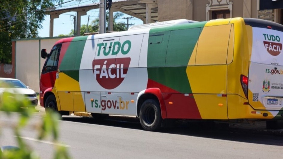 Esta &eacute; uma descri&ccedil;&atilde;o detalhada da imagem para fins de acessibilidade:

Vis&atilde;o Geral
A imagem mostra uma unidade m&oacute;vel de atendimento ao cidad&atilde;o estacionada em uma pra&ccedil;a urbana. O ve&iacute;culo &eacute; um &ocirc;nibus adaptado, personalizado com cores vibrantes e logotipos governamentais, servindo como um posto de servi&ccedil;os itinerante.

Detalhes do Ve&iacute;culo
Design Externo: O &ocirc;nibus &eacute; predominantemente branco, mas apresenta uma cobertura adesiva colorida em tons de azul, laranja, amarelo e verde na parte central e traseira.

Identifica&ccedil;&atilde;o: Na lateral do ve&iacute;culo, destaca-se o logotipo "Tudo F&aacute;cil" em letras grandes, acompanhado da frase "Governo do Estado do Rio Grande do Sul". Tamb&eacute;m &eacute; vis&iacute;vel a marca do programa "RS Digital".

Estrutura: O &ocirc;nibus possui janelas amplas com molduras pretas e uma porta de acesso na parte central. Um toldo retr&aacute;til azul est&aacute; recolhido acima das janelas laterais.

Ambiente e Entorno
Localiza&ccedil;&atilde;o: O ve&iacute;culo est&aacute; posicionado sobre um cal&ccedil;ad&atilde;o de pedras irregulares (estilo basalto) em uma pra&ccedil;a arborizada.

Vegeta&ccedil;&atilde;o e Paisagem: Ao fundo, observam-se &aacute;rvores com folhagens verdes e densas, al&eacute;m de pr&eacute;dios residenciais e comerciais de m&eacute;dio porte sob um c&eacute;u claro.

Mobili&aacute;rio Urbano: Pr&oacute;ximo ao &ocirc;nibus, h&aacute; bancos de pra&ccedil;a de madeira com estrutura de ferro e postes de ilumina&ccedil;&atilde;o p&uacute;blica.

Composi&ccedil;&atilde;o e Ilumina&ccedil;&atilde;o
&Acirc;ngulo: A foto foi tirada de uma perspectiva lateral, mostrando quase toda a extens&atilde;o do &ocirc;nibus, do lado do passageiro.

Luz: A ilumina&ccedil;&atilde;o &eacute; natural e diurna, com sombras suaves projetadas abaixo do ve&iacute;culo, indicando um dia de sol entre nuvens.