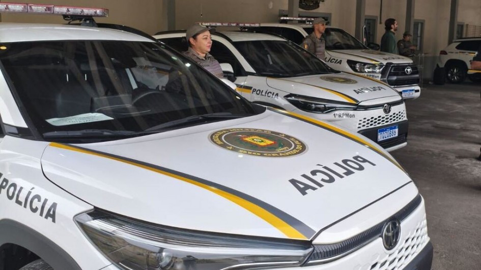 Viaturas da Brigada Militar alinhadas em &aacute;rea coberta, com padroniza&ccedil;&atilde;o visual branca, faixas amarelas e identifica&ccedil;&atilde;o policial. Os ve&iacute;culos est&atilde;o estacionados lado a lado, com ilumina&ccedil;&atilde;o interna do pavilh&atilde;o e estrutura administrativa ao fundo.