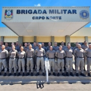 A fotografia colorida mostra um grupo de policiais militares em frente a uma sede da corpora&ccedil;&atilde;o.

Detalhes da cena:

O Grupo: Cerca de 25 policiais (homens e mulheres) est&atilde;o posicionados em forma&ccedil;&atilde;o para uma foto oficial. Eles est&atilde;o organizados em tr&ecirc;s fileiras horizontais, em degraus, todos em p&eacute;, com as m&atilde;os para tr&aacute;s e express&atilde;o s&eacute;ria.

O Uniforme: Todos vestem o uniforme operacional da Brigada Militar do Rio Grande do Sul, composto por camisas e cal&ccedil;as em tom cinza-bandeirante, cintos pretos e botas pretas.

O Pr&eacute;dio: Ao fundo, o grupo est&aacute; posicionado sob a marquise de um pr&eacute;dio de cor clara. Na fachada da marquise, l&ecirc;-se em letras pretas e grandes: "BRIGADA MILITAR", e logo abaixo, em letras menores, "CRPO NORTE". O bras&atilde;o oficial da corpora&ccedil;&atilde;o est&aacute; vis&iacute;vel &agrave; esquerda e &agrave; direita do letreiro.

Ambiente e Ilumina&ccedil;&atilde;o: A foto foi tirada sob luz solar intensa e c&eacute;u azul com poucas nuvens. As sombras est&atilde;o projetadas diretamente abaixo dos policiais, indicando que o sol est&aacute; em uma posi&ccedil;&atilde;o elevada. H&aacute; rampas de acessibilidade com corrim&atilde;os brancos nas laterais do grupo.