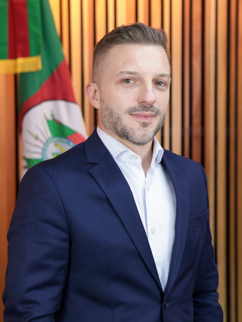 Homem pousa para foto com roupas sociais. Atr&aacute;s dele, uma bandeira do Rio Grande do Sul