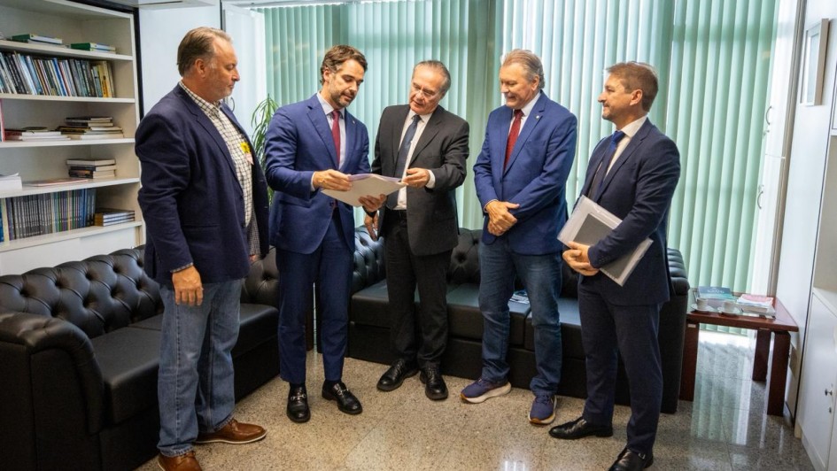 Imagem mostra cinco homens em p&eacute; em uma sala de espera com dois deles trocando documentos.