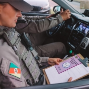 Foto mostra policial militar sentada na carona de uma viatura. Ela est&aacute; com uma prancheta com um panfelto lil&aacute;s. Outro policial est&aacute; sentado ao volante. Dentro do carro, h&aacute; um r&aacute;dio comunicador. 