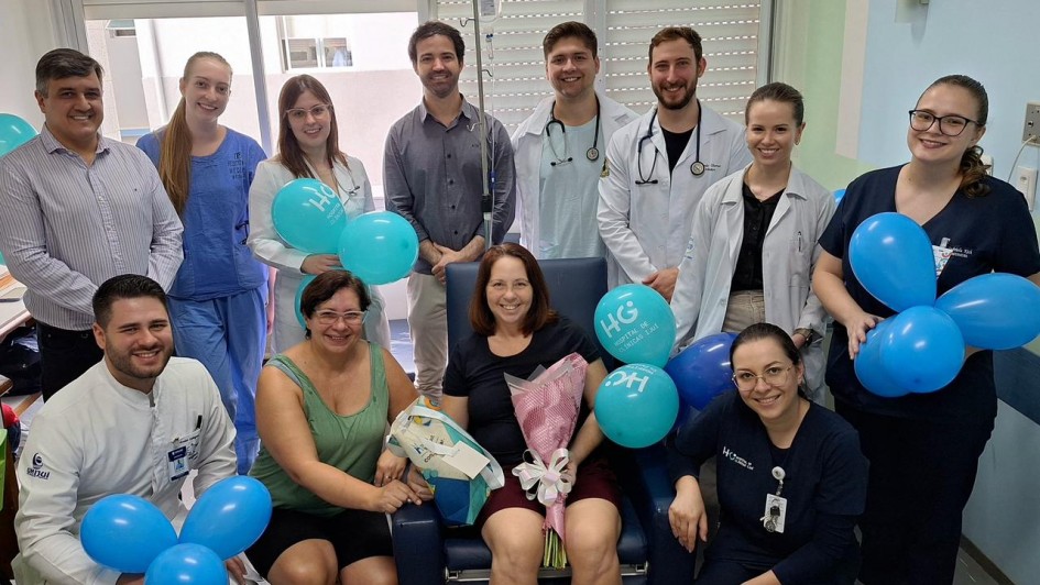 A fotografia colorida registra um momento de celebra&ccedil;&atilde;o em um ambiente hospitalar. Um grupo de doze pessoas rodeia uma mulher sentada ao centro, que sorri enquanto segura um buqu&ecirc; de flores.

Detalhes da cena:

A paciente: Uma mulher de cabelos castanhos claros est&aacute; sentada em uma poltrona hospitalar azul. Ela segura um buqu&ecirc; de flores envolto em papel rosa e uma sacola de presente.

A equipe de sa&uacute;de e acompanhantes: Ao redor dela, dez profissionais de sa&uacute;de e um homem em trajes civis posam para a foto. Os profissionais vestem jalecos brancos com estetosc&oacute;pios no pesco&ccedil;o ou uniformes privativos (scrubs) em tons de azul claro e azul marinho. Todos sorriem para a c&acirc;mera.

Decora&ccedil;&atilde;o festiva: O grupo segura diversos bal&otilde;es azuis inflados. Alguns bal&otilde;es possuem o logotipo "HCI - Hospital de Cl&iacute;nicas Iju&iacute;" impresso em branco.

O ambiente: A cena ocorre dentro de um quarto de hospital. Ao fundo, v&ecirc;-se uma janela com persianas horizontais fechadas e um suporte de soro com uma bolsa de fluido pendurada, conectada &agrave; paciente.

Ilumina&ccedil;&atilde;o: A luz &eacute; clara e uniforme, caracter&iacute;stica de ambientes cl&iacute;nicos, transmitindo uma atmosfera de al&iacute;vio e comemora&ccedil;&atilde;o.