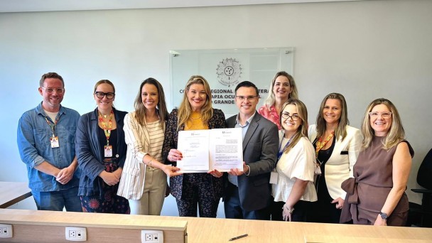 A fotografia colorida mostra um grupo de nove pessoas (tr&ecirc;s homens e seis mulheres) em p&eacute;, posicionadas lado a lado em um ambiente de escrit&oacute;rio, celebrando a assinatura de um acordo.

Detalhes da cena:

O ato central: No centro do grupo, uma mulher loira vestindo um blazer estampado e um homem de &oacute;culos vestindo um palet&oacute; cinza seguram juntos um documento oficial aberto. Eles sorriem para a c&acirc;mera, simbolizando a formaliza&ccedil;&atilde;o de uma parceria. O documento exibe o bras&atilde;o do Estado do Rio Grande do Sul no topo.

Os participantes: As outras sete pessoas est&atilde;o distribu&iacute;das ao redor do casal central. Elas vestem trajes que variam entre o casual e o formal (blazers, camisas sociais e blusas coloridas). V&aacute;rias pessoas no grupo utilizam crach&aacute;s com cord&otilde;es vermelhos e amarelos pendurados no pesco&ccedil;o.

O ambiente: O grupo est&aacute; atr&aacute;s de uma bancada de madeira clara com tomadas embutidas. Ao fundo, em uma parede branca, h&aacute; uma placa de acr&iacute;lico transparente com um bras&atilde;o circular e o texto: "CONSELHO REGIONAL DE FISIOTERAPIA E TERAPIA OCUPACIONAL DO RIO GRANDE DO SUL".

Ilumina&ccedil;&atilde;o: A foto possui ilumina&ccedil;&atilde;o frontal clara e sombras suaves, com um clima profissional e positivo. Uma caneta preta est&aacute; apoiada sobre a bancada de madeira, em frente ao grupo.