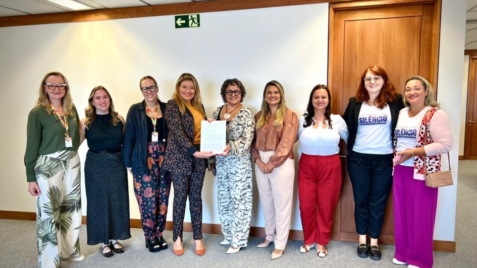 A fotografia colorida mostra um grupo de nove mulheres em p&eacute;, posicionadas lado a lado em um ambiente de escrit&oacute;rio com paredes brancas e detalhes em madeira. Elas celebram a entrega de um documento oficial.

Detalhes da cena:

O ato central: No centro do grupo, duas mulheres seguram juntas uma folha de papel branca com texto impresso. A mulher &agrave; esquerda veste um conjunto de blazer e cal&ccedil;a com estampa escura e detalhes laranjas; a mulher &agrave; direita veste um conjunto branco com estampas pretas delicadas e &oacute;culos de arma&ccedil;&atilde;o escura. Ambas sorriem para a c&acirc;mera.

As participantes: As outras sete mulheres est&atilde;o distribu&iacute;das lateralmente. Elas vestem roupas variadas, incluindo blusas em tons de verde, preto, rosa e branco, al&eacute;m de cal&ccedil;as coloridas (vermelha, magenta e estampadas). Duas mulheres &agrave; direita vestem camisetas brancas com a palavra "SIL&Ecirc;NCIO" em destaque, parte de uma campanha institucional. Algumas participantes usam crach&aacute;s com cord&otilde;es coloridos no pesco&ccedil;o.

O ambiente: O grupo est&aacute; em um corredor ou sala ampla com carpete cinza. Ao fundo, h&aacute; uma parede branca com uma placa de sinaliza&ccedil;&atilde;o de sa&iacute;da (fundo verde com um boneco branco correndo) e uma porta de madeira fechada &agrave; direita.

Ilumina&ccedil;&atilde;o: A luz &eacute; artificial, vinda do teto, criando uma ilumina&ccedil;&atilde;o clara e uniforme sobre todas as pessoas, transmitindo um tom de formalidade e coopera&ccedil;&atilde;o.