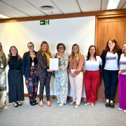 A fotografia colorida mostra um grupo de nove mulheres em p&eacute;, posicionadas lado a lado em um ambiente de escrit&oacute;rio com paredes brancas e detalhes em madeira. Elas celebram a entrega de um documento oficial.

Detalhes da cena:

O ato central: No centro do grupo, duas mulheres seguram juntas uma folha de papel branca com texto impresso. A mulher &agrave; esquerda veste um conjunto de blazer e cal&ccedil;a com estampa escura e detalhes laranjas; a mulher &agrave; direita veste um conjunto branco com estampas pretas delicadas e &oacute;culos de arma&ccedil;&atilde;o escura. Ambas sorriem para a c&acirc;mera.

As participantes: As outras sete mulheres est&atilde;o distribu&iacute;das lateralmente. Elas vestem roupas variadas, incluindo blusas em tons de verde, preto, rosa e branco, al&eacute;m de cal&ccedil;as coloridas (vermelha, magenta e estampadas). Duas mulheres &agrave; direita vestem camisetas brancas com a palavra "SIL&Ecirc;NCIO" em destaque, parte de uma campanha institucional. Algumas participantes usam crach&aacute;s com cord&otilde;es coloridos no pesco&ccedil;o.

O ambiente: O grupo est&aacute; em um corredor ou sala ampla com carpete cinza. Ao fundo, h&aacute; uma parede branca com uma placa de sinaliza&ccedil;&atilde;o de sa&iacute;da (fundo verde com um boneco branco correndo) e uma porta de madeira fechada &agrave; direita.

Ilumina&ccedil;&atilde;o: A luz &eacute; artificial, vinda do teto, criando uma ilumina&ccedil;&atilde;o clara e uniforme sobre todas as pessoas, transmitindo um tom de formalidade e coopera&ccedil;&atilde;o.