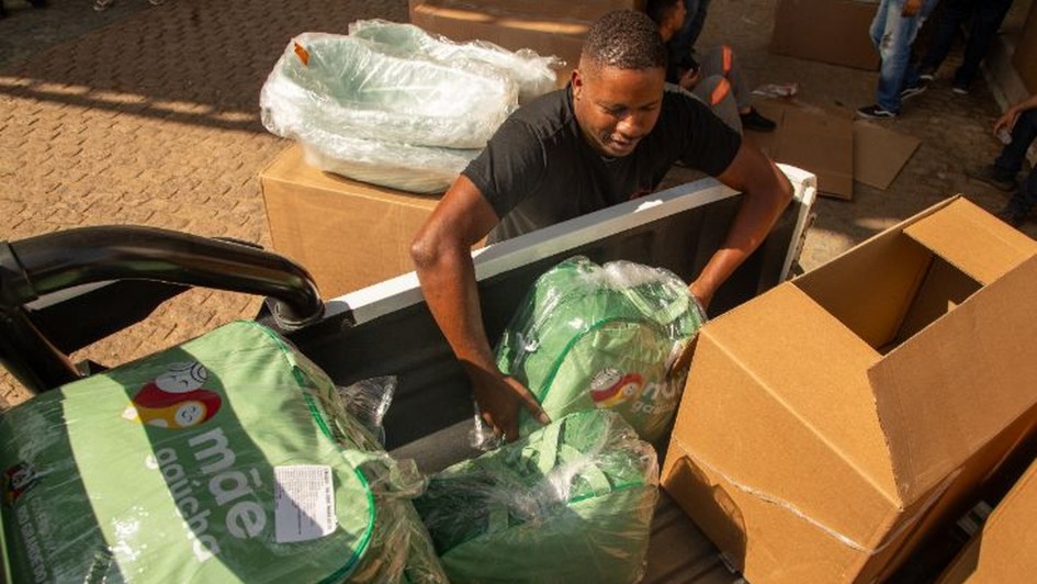 Foto mostra um servidor carregando uma carga em um ve&iacute;culo com itens do kit do M&atilde;e Ga&uacute;cha, com sacolas em verde claro e caixas de papel&atilde;o. 