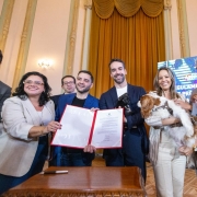 Foto mostra o governador Eduardo Leite segurando um pequeno cachorro preto e o vice Gabriel Leite, junto com a secret&aacute;ria que seguram um documento assinado. Outra mulher segura um cachorro branco e manchas marrons. Outras autoridades est&atilde;o juntos e aplaudem a cena. 