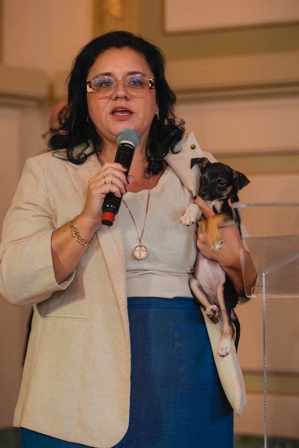 Foto mostra secretária Marjorie Kauffmann discursando em pé, com o microfone em uma mão, e na outra abraça um filhote de cachorro, de pelagem preta. 