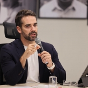 Esta &eacute; uma descri&ccedil;&atilde;o da imagem com foco em acessibilidade:

A fotografia colorida mostra um plano m&eacute;dio do governador Eduardo Leite durante uma atividade oficial de trabalho.

Detalhes da cena:

O orador: Eduardo Leite, um homem branco com barba e cabelos castanhos curtos, est&aacute; sentado &agrave; mesa. Ele veste um palet&oacute; azul-marinho sobre uma camiseta branca. Com a m&atilde;o esquerda, ele segura um microfone prateado pr&oacute;ximo &agrave; boca, enquanto a m&atilde;o direita gesticula, segurando uma caneta branca, indicando que est&aacute; em meio a uma explica&ccedil;&atilde;o. Ele usa um rel&oacute;gio de pulso com pulseira clara.

A mesa de trabalho: &Agrave; sua frente, sobre a mesa de tom claro, h&aacute; um tablet (iPad) apoiado em uma capa-teclado preta, pap&eacute;is de trabalho e um copo de vidro com &aacute;gua.

O ambiente: O fundo &eacute; composto por uma parede de cor neutra (cinza claro). Atr&aacute;s do governador, &eacute; poss&iacute;vel ver parte de fotografias em preto e branco emolduradas, que decoram o ambiente da reuni&atilde;o.

Composi&ccedil;&atilde;o e Ilumina&ccedil;&atilde;o: A imagem tem um enquadramento focado no governador, com o fundo levemente desfocado para dar destaque &agrave; sua express&atilde;o e fala. A
