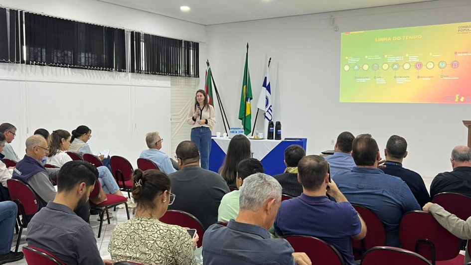 Foto mostra assembleia da Consulta Popular com uma mulher falando em p&eacute; diante da plateia.