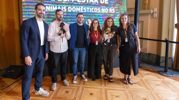 A fotografia colorida registra um momento de intera&ccedil;&atilde;o entre autoridades e a comunidade durante um evento oficial voltado ao bem-estar animal.

Detalhes da cena:

Os participantes: No centro da imagem, o Governador Eduardo Leite, vestindo uma camisa social azul clara e cal&ccedil;a escura, est&aacute; levemente agachado para interagir com um cachorro. &Agrave; esquerda, uma mulher de blusa rosa e &oacute;culos sorri enquanto observa a cena. Ao redor deles, outras pessoas acompanham o momento em um ambiente de celebra&ccedil;&atilde;o.

O animal: Um cachorro de m&eacute;dio porte, com pelagem curta e amarelada (semelhante a um Labrador ou vira-lata), est&aacute; em p&eacute; &agrave; frente do governador. O animal parece calmo e recebe aten&ccedil;&atilde;o das autoridades.

O ambiente: O evento ocorre em um espa&ccedil;o amplo com ilumina&ccedil;&atilde;o natural. Ao fundo, &eacute; poss&iacute;vel ver estruturas que sugerem um parque ou &aacute;rea externa coberta, com &aacute;rvores e folhagens vis&iacute;veis ao longe. H&aacute; tamb&eacute;m um banner ou painel com identidades visuais do governo do Estado.

Composi&ccedil;&atilde;o e Ilumina&ccedil;&atilde;o: A foto captura o grupo da cintura para cima, com foco central na intera&ccedil;&atilde;o entre o governador e o c&atilde;o. A luz do dia &eacute; clara, destacando as express&otilde;es de satisfa&ccedil;&atilde;o dos presentes e refor&ccedil;ando o car&aacute;ter p&uacute;blico e amig&aacute;vel da agenda.