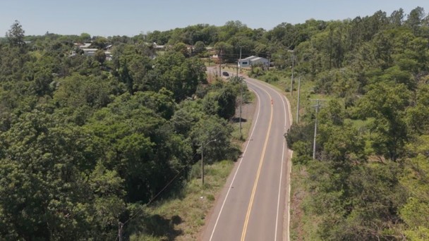 A fotografia colorida, tirada de um &acirc;ngulo elevado (perspectiva a&eacute;rea), mostra um trecho sinuoso de uma rodovia rec&eacute;m-asfaltada que atravessa uma &aacute;rea rural e montanhosa.

Detalhes da cena:

A rodovia: O asfalto &eacute; de um tom cinza escuro intenso, indicando pavimenta&ccedil;&atilde;o nova. A estrada serpenteia pela paisagem, apresentando curvas suaves. A sinaliza&ccedil;&atilde;o horizontal &eacute; composta por faixas brancas cont&iacute;nuas nas bordas e uma faixa amarela cont&iacute;nua no centro, dividindo as pistas.

A paisagem: Ao redor da rodovia, a vegeta&ccedil;&atilde;o &eacute; densa e predominantemente verde, composta por &aacute;rvores nativas, arbustos e &aacute;reas de campo. O terreno &eacute; acidentado, com eleva&ccedil;&otilde;es que formam vales por onde a estrada foi constru&iacute;da.

O horizonte: Ao fundo, veem-se sucessivas camadas de morros e montanhas cobertos por vegeta&ccedil;&atilde;o, que se tornam mais claros e azulados devido &agrave; dist&acirc;ncia. O c&eacute;u &eacute; de um tom azul p&aacute;lido, quase branco, sugerindo um dia claro com luz solar difusa.

Composi&ccedil;&atilde;o e Ilumina&ccedil;&atilde;o: A luz natural destaca o contraste entre o cinza escuro do asfalto novo e o verde vibrante da natureza ao redor. A imagem transmite uma sensa&ccedil;&atilde;o de moderniza&ccedil;&atilde;o da infraestrutura integrada ao meio ambiente.