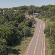 A fotografia colorida, tirada de um &acirc;ngulo elevado (perspectiva a&eacute;rea), mostra um trecho sinuoso de uma rodovia rec&eacute;m-asfaltada que atravessa uma &aacute;rea rural e montanhosa.

Detalhes da cena:

A rodovia: O asfalto &eacute; de um tom cinza escuro intenso, indicando pavimenta&ccedil;&atilde;o nova. A estrada serpenteia pela paisagem, apresentando curvas suaves. A sinaliza&ccedil;&atilde;o horizontal &eacute; composta por faixas brancas cont&iacute;nuas nas bordas e uma faixa amarela cont&iacute;nua no centro, dividindo as pistas.

A paisagem: Ao redor da rodovia, a vegeta&ccedil;&atilde;o &eacute; densa e predominantemente verde, composta por &aacute;rvores nativas, arbustos e &aacute;reas de campo. O terreno &eacute; acidentado, com eleva&ccedil;&otilde;es que formam vales por onde a estrada foi constru&iacute;da.

O horizonte: Ao fundo, veem-se sucessivas camadas de morros e montanhas cobertos por vegeta&ccedil;&atilde;o, que se tornam mais claros e azulados devido &agrave; dist&acirc;ncia. O c&eacute;u &eacute; de um tom azul p&aacute;lido, quase branco, sugerindo um dia claro com luz solar difusa.

Composi&ccedil;&atilde;o e Ilumina&ccedil;&atilde;o: A luz natural destaca o contraste entre o cinza escuro do asfalto novo e o verde vibrante da natureza ao redor. A imagem transmite uma sensa&ccedil;&atilde;o de moderniza&ccedil;&atilde;o da infraestrutura integrada ao meio ambiente.