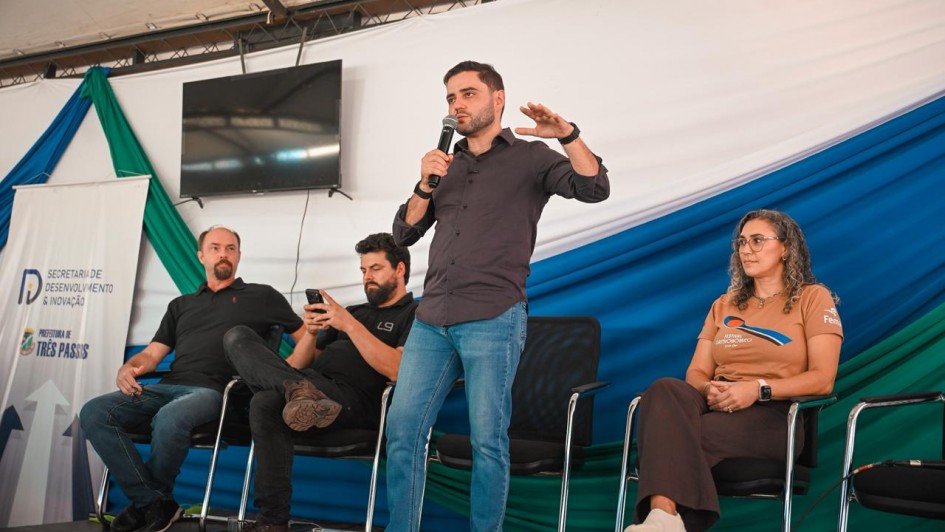 Quatro pessoasl em um palco, tr&ecirc;s sentadas e uma em p&eacute; falando ao microfone para pessoas &agrave; frente que n&atilde;o aparecem na foto