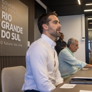 Dois homens sentados com os bra&ccedil;os apoiados em uma grande mesa, &agrave; frente de um painel com os dizeres: Governo do Estado do Rio Grande do Sul O Futuro nos Une. Um terceiro homem em p&eacute; olha para ambos 